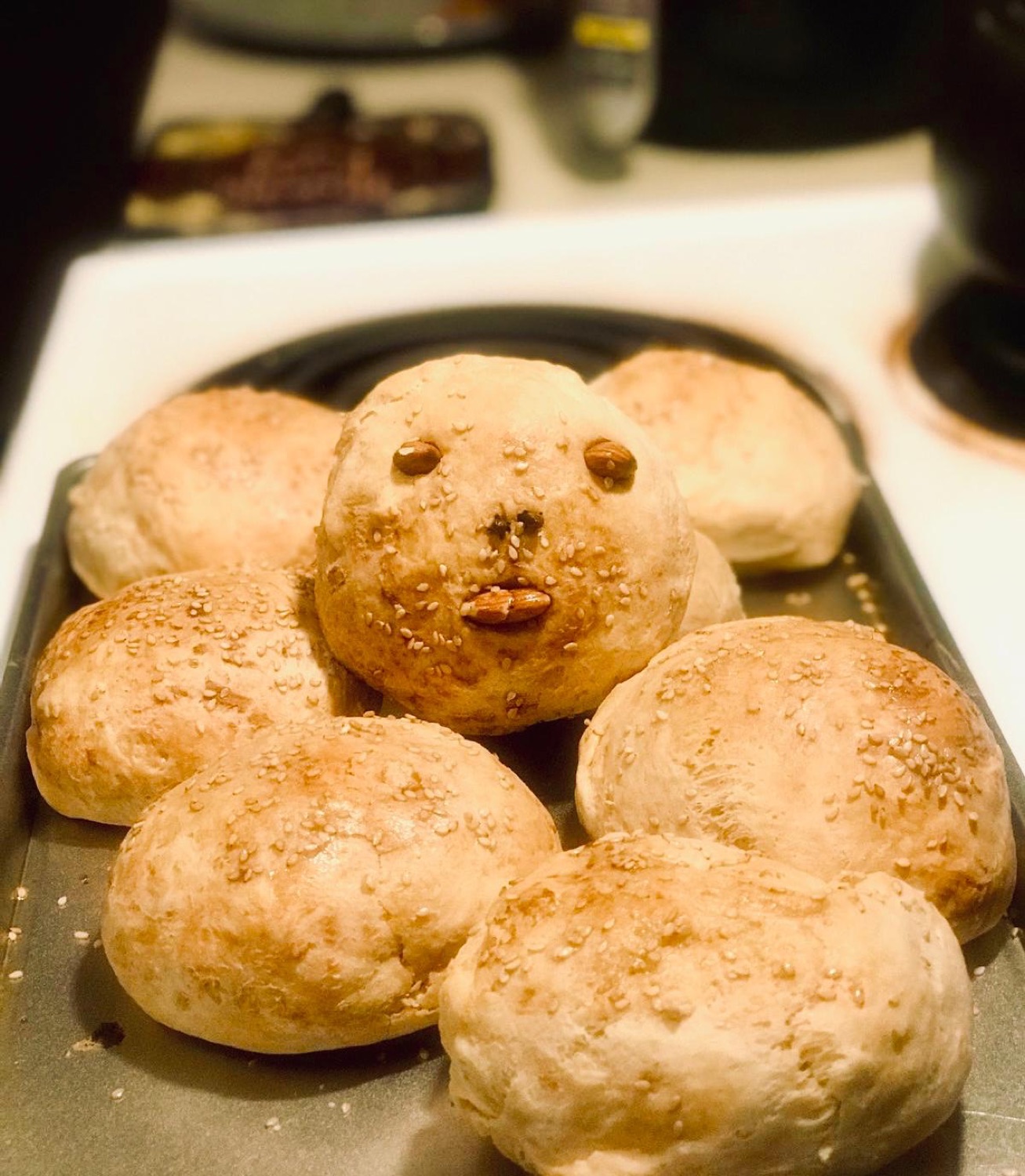 A tray of golden sesame-topped bread rolls, one with an almond face