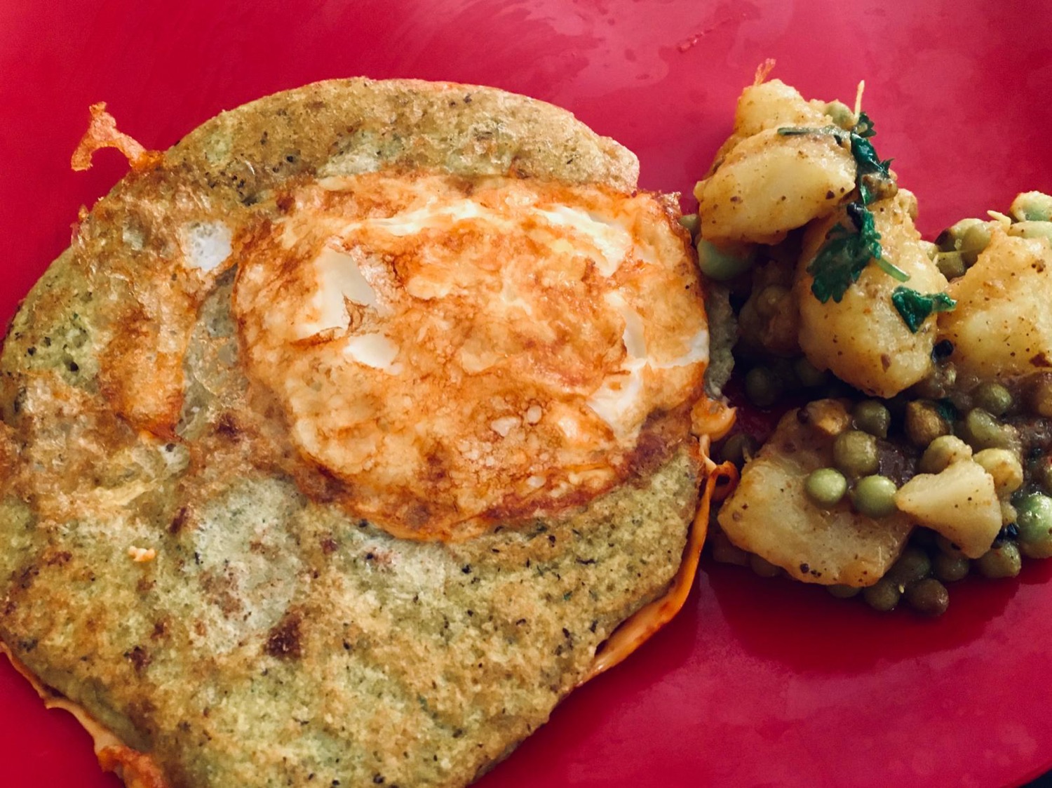 A green herb pancake topped with a fried egg, with potato and pea curry on a red plate