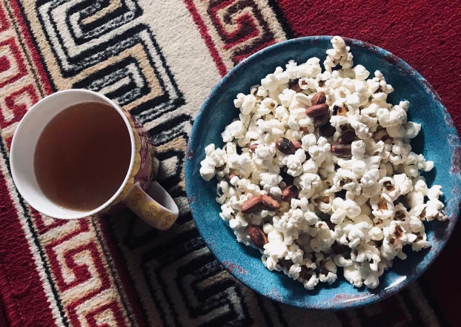 A bowl of popcorn with roasted peanuts beside a cup of milk tea on a patterned carpet