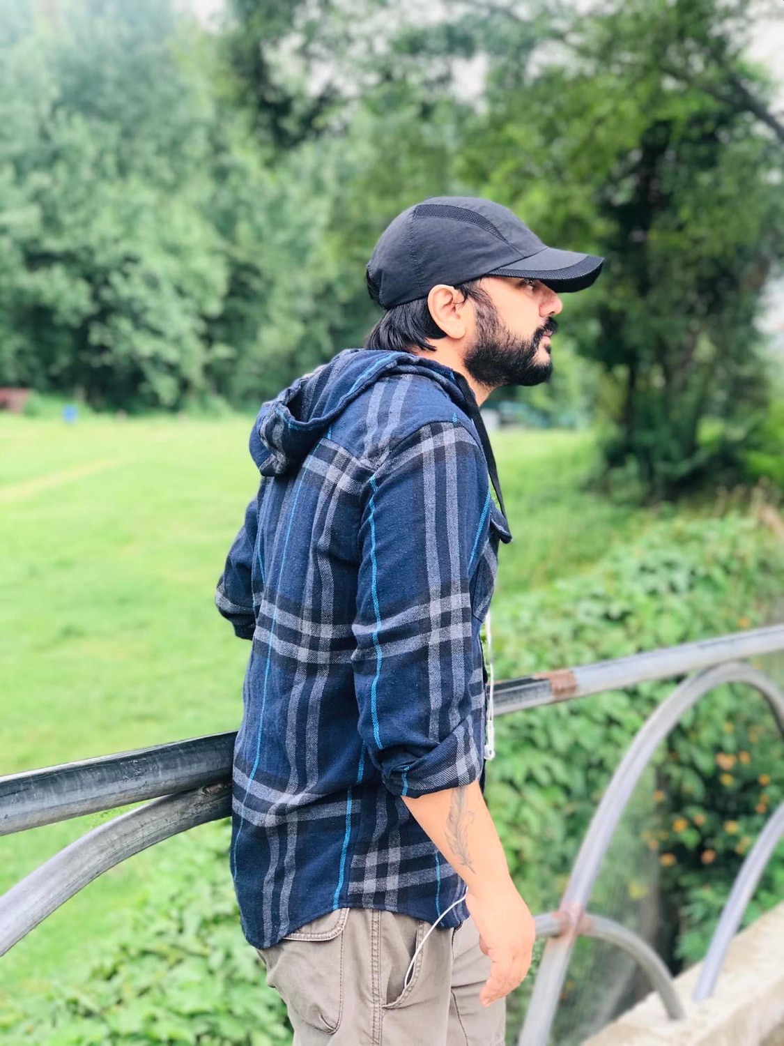 Udeep in a cap and flannel shirt, leaning on a bridge railing above a green field