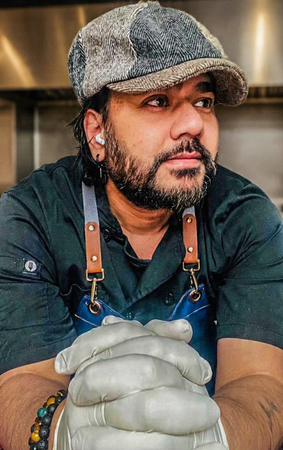 Udeep in a herringbone flat cap and denim apron, gloved hands folded, in a restaurant kitchen