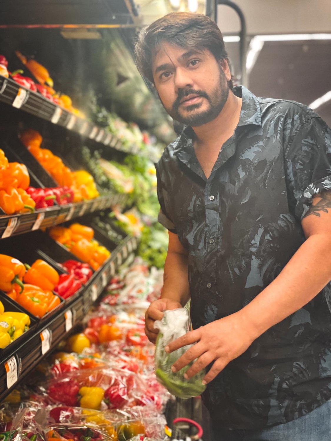 Udeep in the produce aisle holding a bag of green chilies, surrounded by bell peppers